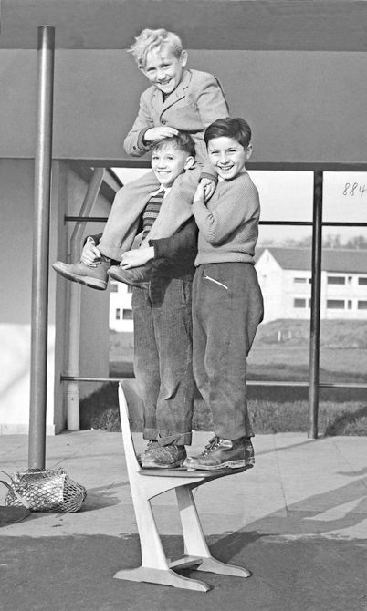 Black and white image taken 1950 of three children standing all together on a wooden skid chair by VS.