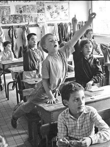 A black and white photo of a classroom focussing on a boy eagerly raising his hand