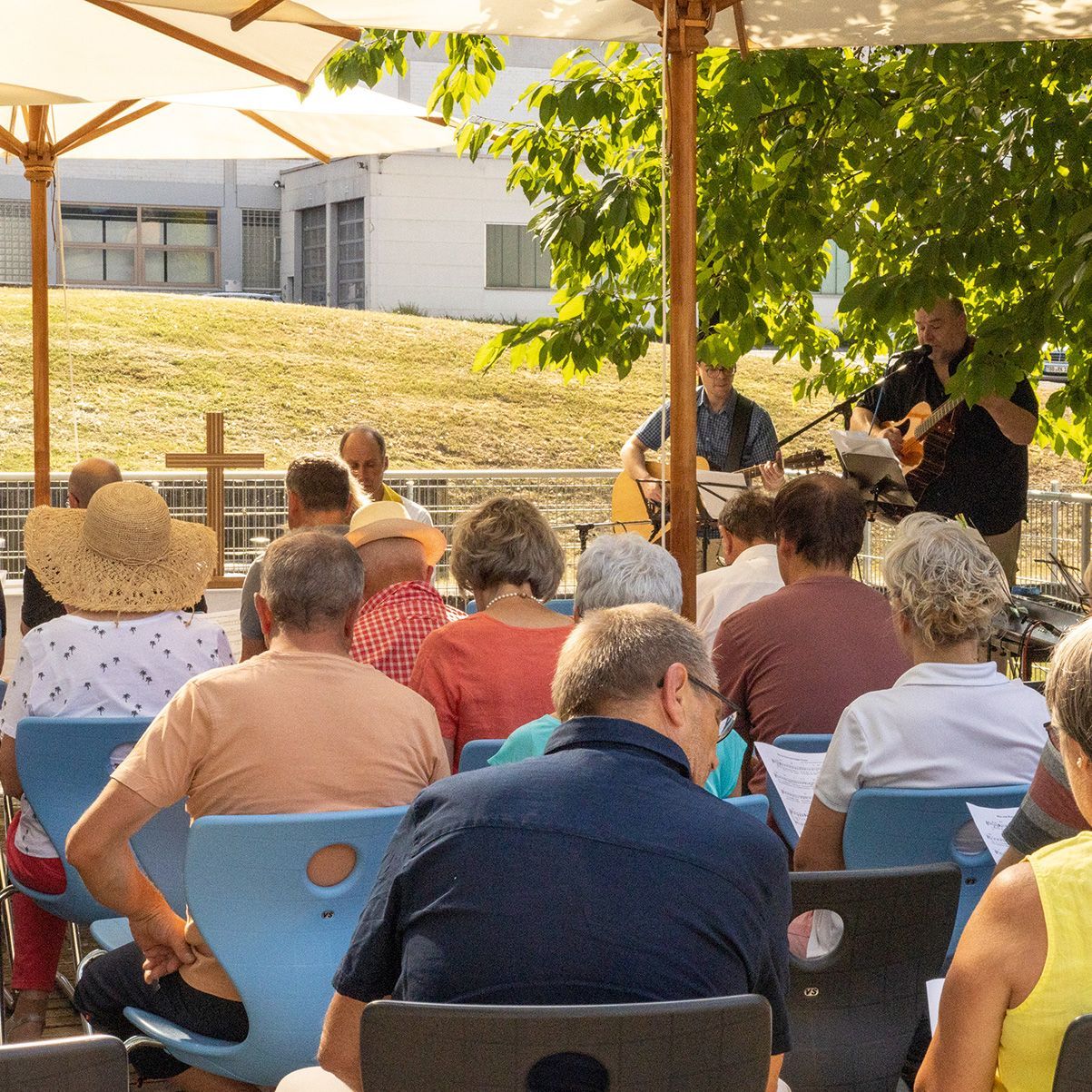 Menschen sitzen draußen auf VS-Stühlen vor einem Kreuz bei einem Gottesdienst