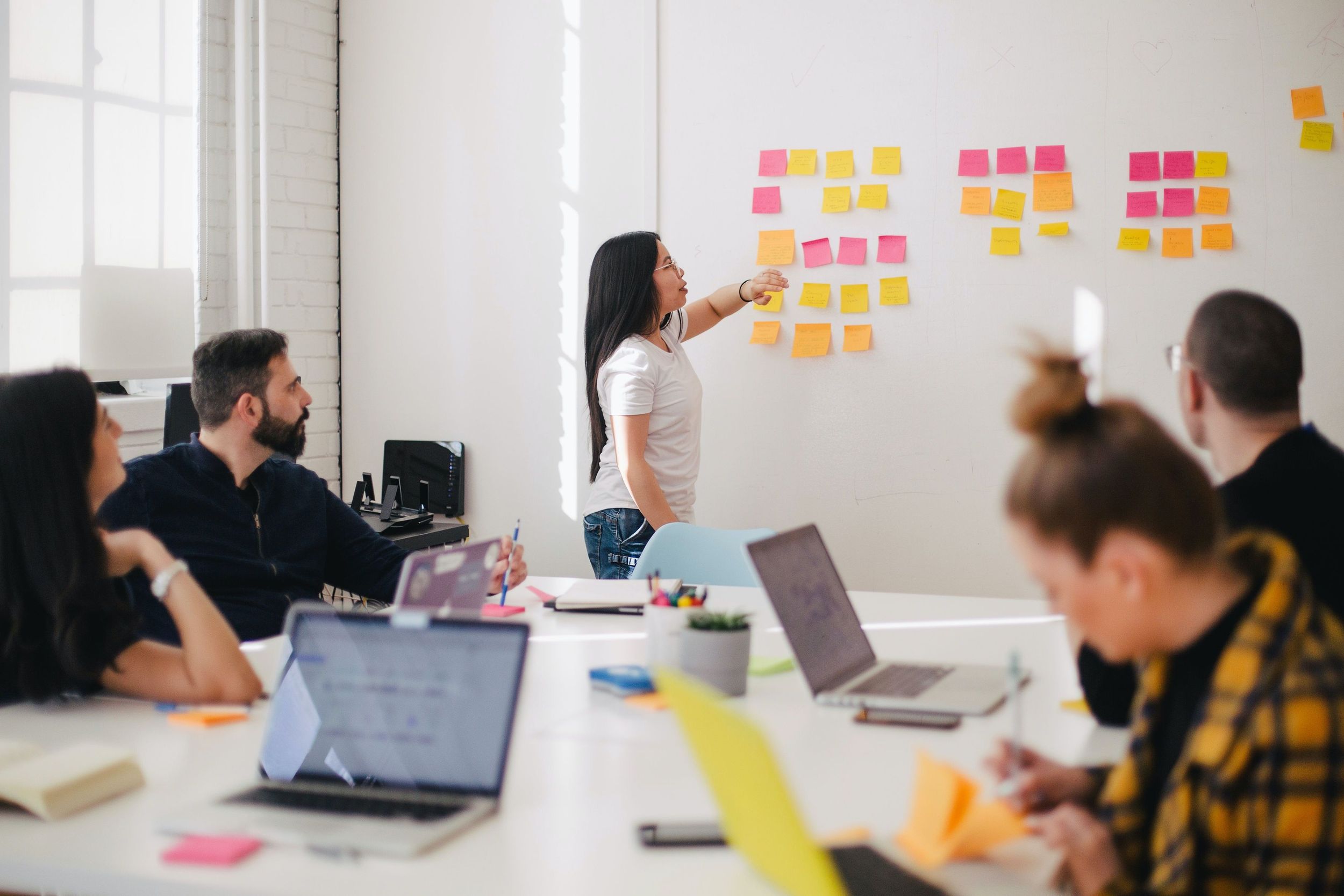 Mehrere Personen in einem Meeting-Raum mit Laptops, hören einer Frau zu, die etwas an einem Whiteboard erklärt