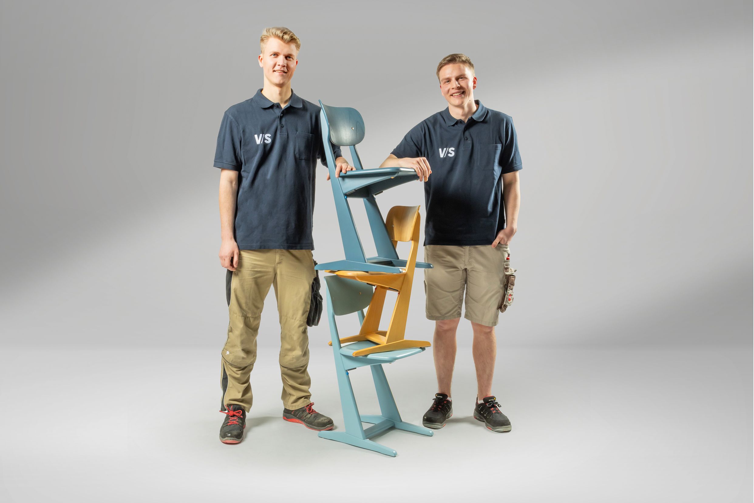 Two young wood technology students with stacked colourful wooden skid chairs from VS laugh into the camera.