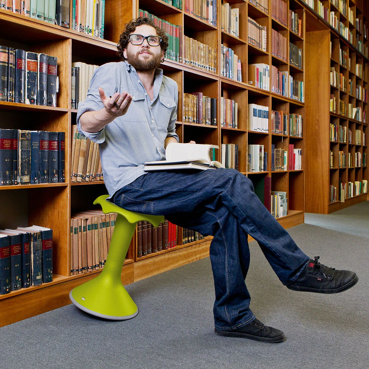 A man holding books sits on a light green Hokki in a library