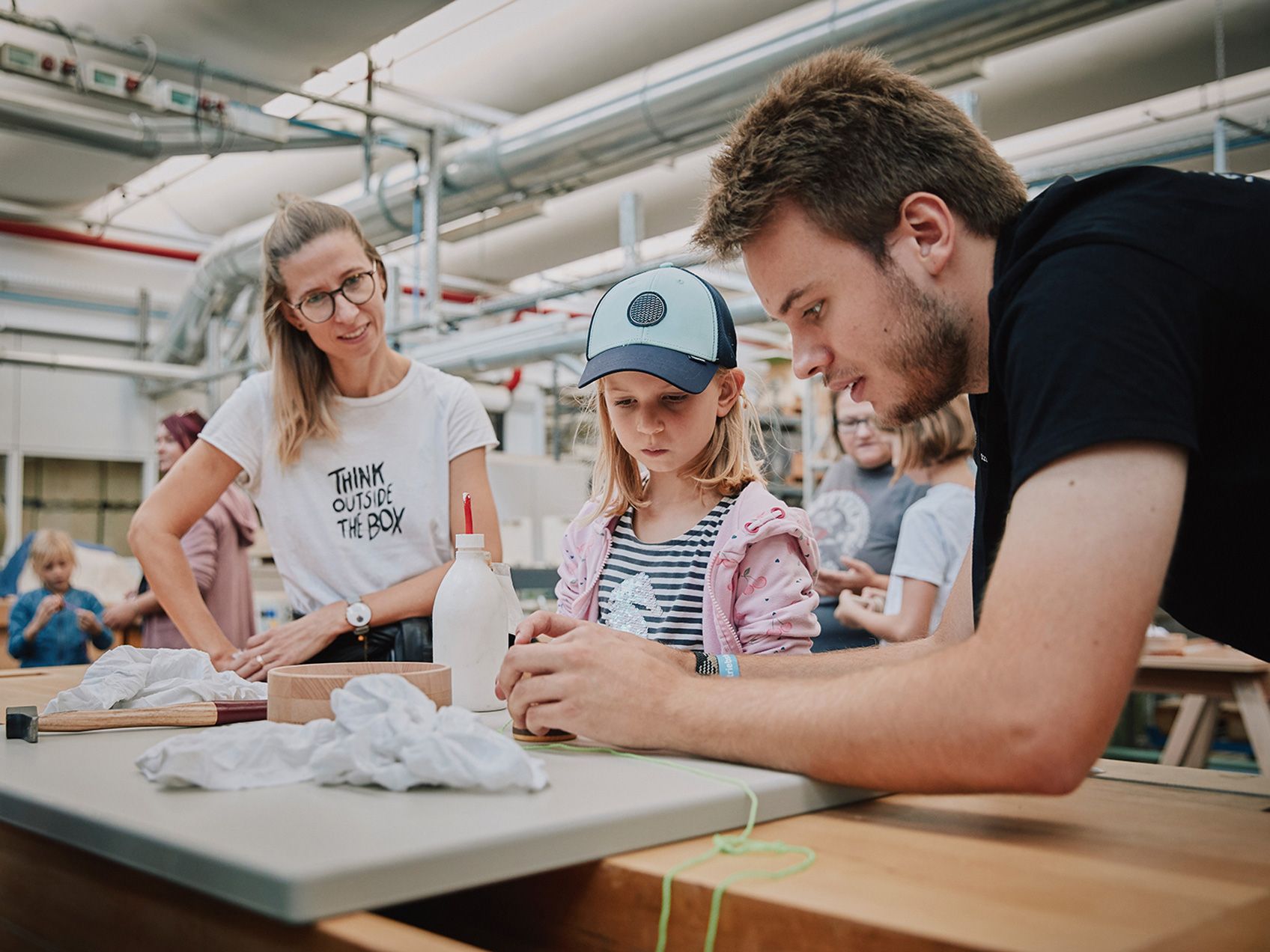Ein Kind mit seiner Mutter und ein Mitarbeiter von VS stehen an einer Werkbank in der Holzwerkstatt und basteln Spielzeug beim Tag der Offenen Tür bei VS.