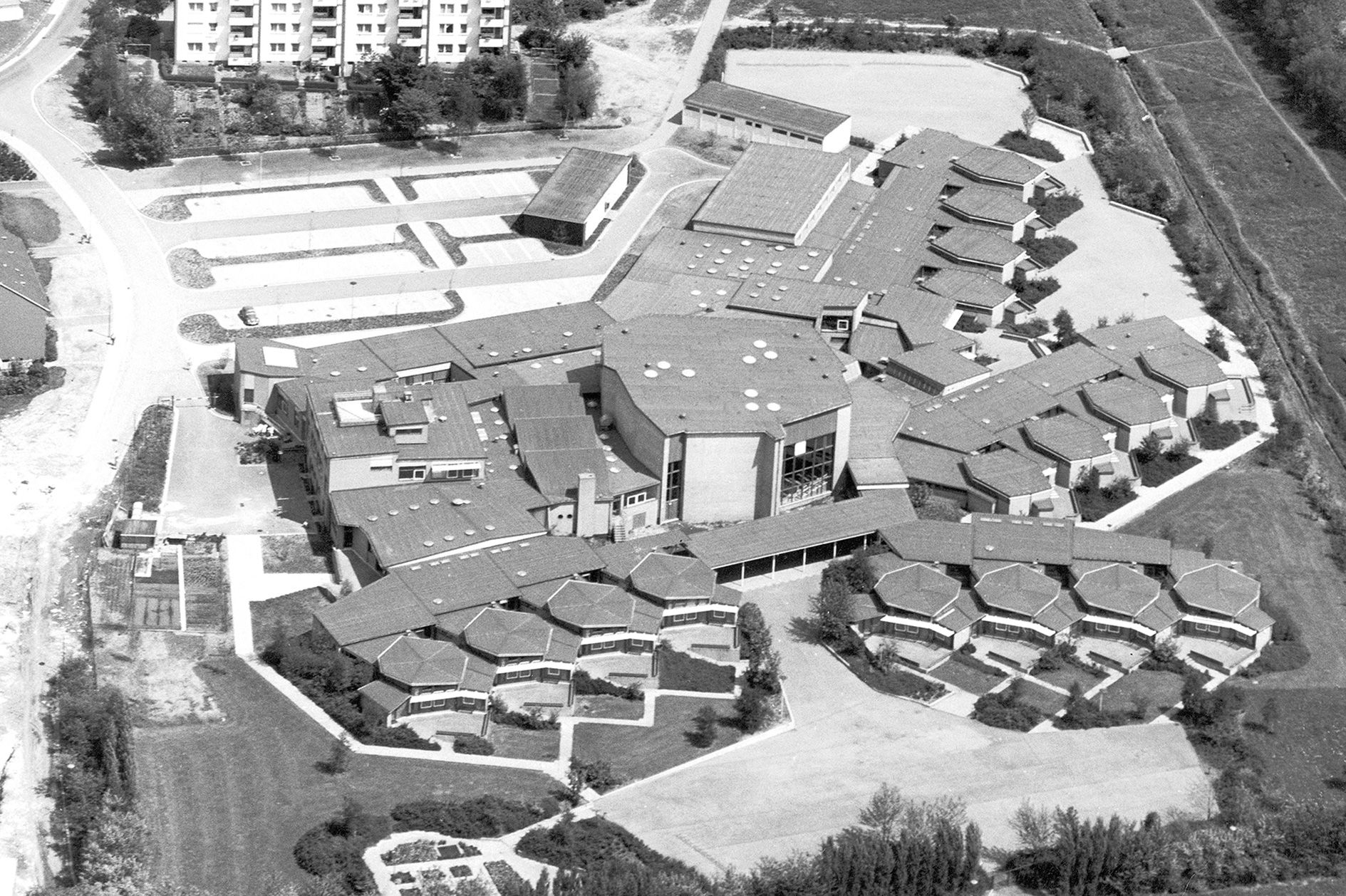 Schwarz-Weiß-Bild aus den 1970ern der von Hans Sharoun entworfenen Grund- und Hauptschule in Marl in Westfalen mit wabenförmigen, einstöckigen Gebäuden um die zentral gelegene, höhere Aula herum