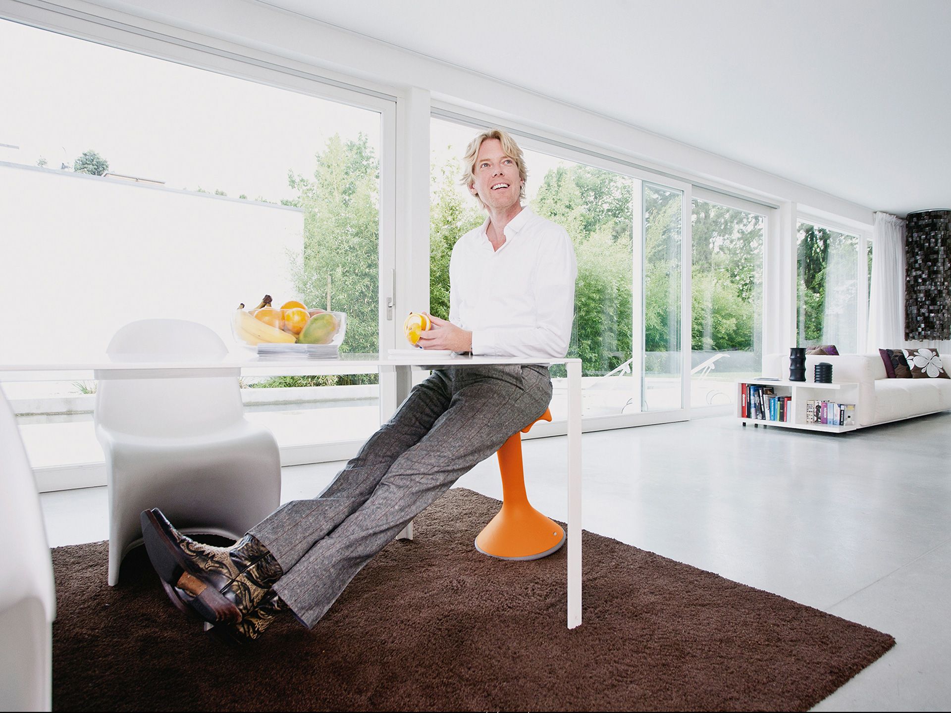 Un homme d'âge moyen est assis à une table sur le tabouret orange mobile Hokki de VS dans une pièce au décor lumineux.