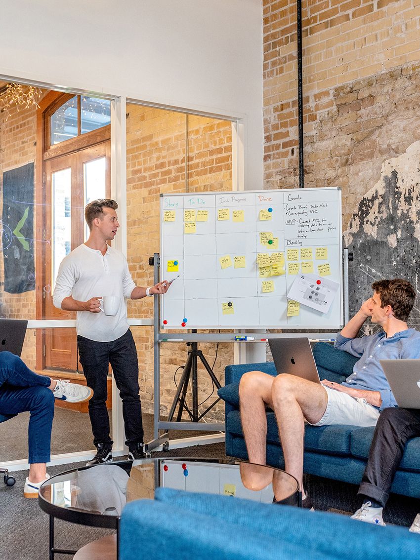 Eine Personen steht vor einem Whiteboard und unterhält sich mit einer weiteren Person, die daneben auf einem Sofa sitzt.