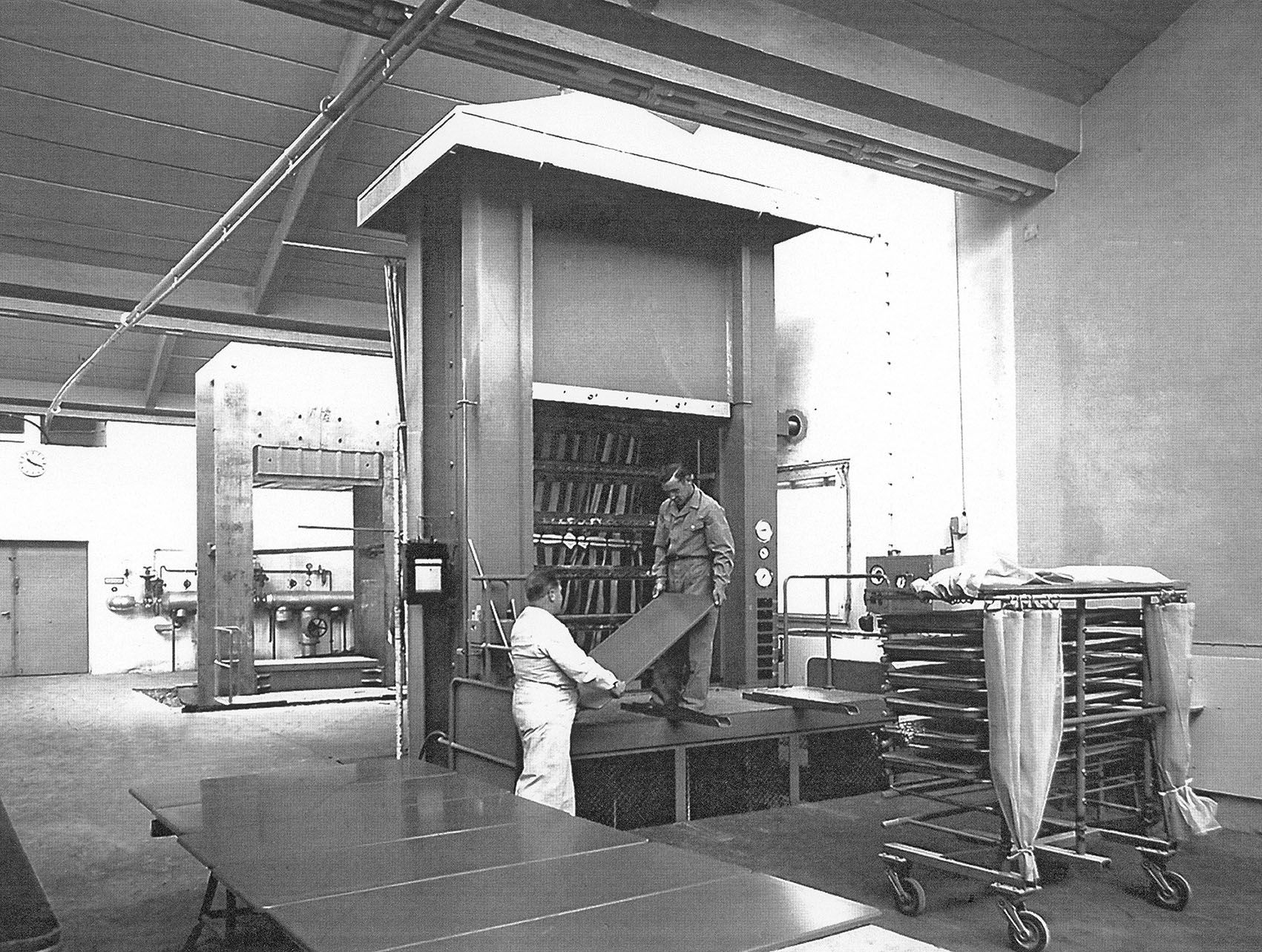 Black and white photo from a factory hall of VS in which two employees are taking a table top out of a Lignodur press.